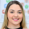 Imagem de Lorena, aluna do curso de fisioterapia no campus Presidente Prudente, trajando jaleco escuro. O fundo é claro e bem iluminado, com equipamentos médicos ao fundo, indicando um laboratório ou sala de práticas acadêmicas na área da saúde.