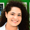 Imagem de Maria, aluna do curso de fisioterapia no campus Presidente Prudente, usando jaleco branco. O fundo apresenta modelos anatômicos em prateleiras, sugerindo um ambiente acadêmico técnico voltado para o estudo prático e científico.