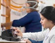 Estudante de Fisioterapia opera aparelho de ventilação mecânica enquanto paciente utiliza máscara respiratória. Ao fundo, a paciente segura barras de apoio em ambiente clínico, demonstrando prática de reabilitação respiratória e motora, com equipamentos modernos disponíveis.