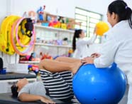 Criança deitada com pernas apoiadas em bola suíça azul realiza exercício supervisionado por estudante de Fisioterapia. Ao fundo, outra aluna auxilia paciente com bola amarela. Sala repleta de brinquedos e materiais pedagógicos, típica de clínica de fisioterapia pediátrica.