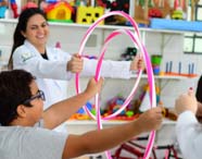 Em sala de aula, estudante de Fisioterapia orienta crianças no uso de bambolês rosa e branco para exercícios de coordenação motora. Prateleiras ao fundo exibem brinquedos educativos, reforçando o ambiente voltado à reabilitação pediátrica e aprendizagem motora.