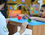 Estudante de Fisioterapia acompanha criança durante atividade de motricidade fina, usando bolas sensoriais vermelha e verde sobre mesa azul. O cenário é colorido, com arcos e painéis decorativos, ressaltando o foco em atendimento didático e desenvolvimento infantil.