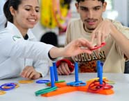 Estudante de Fisioterapia auxilia criança em atividade de encaixe de argolas coloridas em pinos, estimulando habilidades motoras. Mesa branca com diversos acessórios terapêuticos e ambiente vibrante, ideal para reabilitação pediátrica e inclusão lúdica.