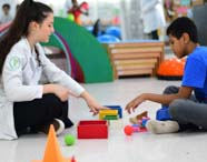 Sentados no chão, estudante de Fisioterapia e criança interagem com blocos coloridos e bolas sensoriais. O espaço possui obstáculos e equipamentos de psicomotricidade, promovendo aprendizado e reabilitação motora em ambiente acolhedor e educativo.