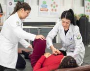 Duas estudantes de Fisioterapia realizam atendimento prático com paciente deitada em colchonete, auxiliando na movimentação das pernas. A sala possui barras paralelas e cartazes explicativos nas paredes, reforçando o caráter acadêmico e clínico do curso.
