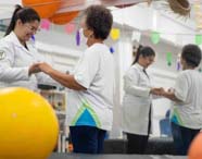 Estudante de Fisioterapia atende idosa em sala decorada, segurando suas mãos para exercício de equilíbrio diante de espelho. Bola grande amarela em destaque, bandeirolas coloridas no teto e reflexo dos participantes ampliam o ambiente de reabilitação humanizada.