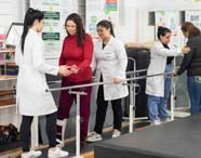 Paciente com máscara respiratória realiza exercício de fortalecimento segurando barras fixas, enquanto estudante de Fisioterapia apoia suas costas. Ambiente equipado com argolas coloridas e outros materiais, típico de clínica universitária de reabilitação motora e respiratória.