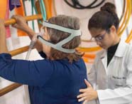 Três estudantes de Fisioterapia avaliam paciente sentada, que utiliza máscara de ventilação não invasiva. Uma aluna estabiliza a paciente, outra apresenta estímulo visual, enquanto a terceira observa. Sala decorada com argolas e bolas, evidenciando ambiente clínico acadêmico.