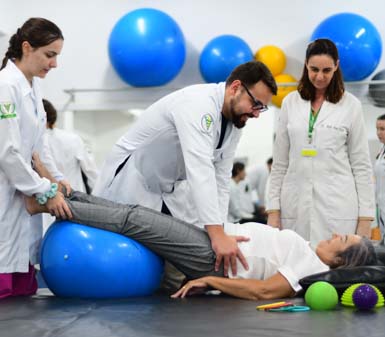 Em sala de aula prática do curso de Fisioterapia, estudantes com jaleco branco auxiliam paciente deitada sobre colchonete preto, realizando exercício com bola suíça azul sob as pernas. Há bolas coloridas ao fundo e acessórios terapêuticos próximos, destacando ambiente moderno e recursos da instituição de ensino.