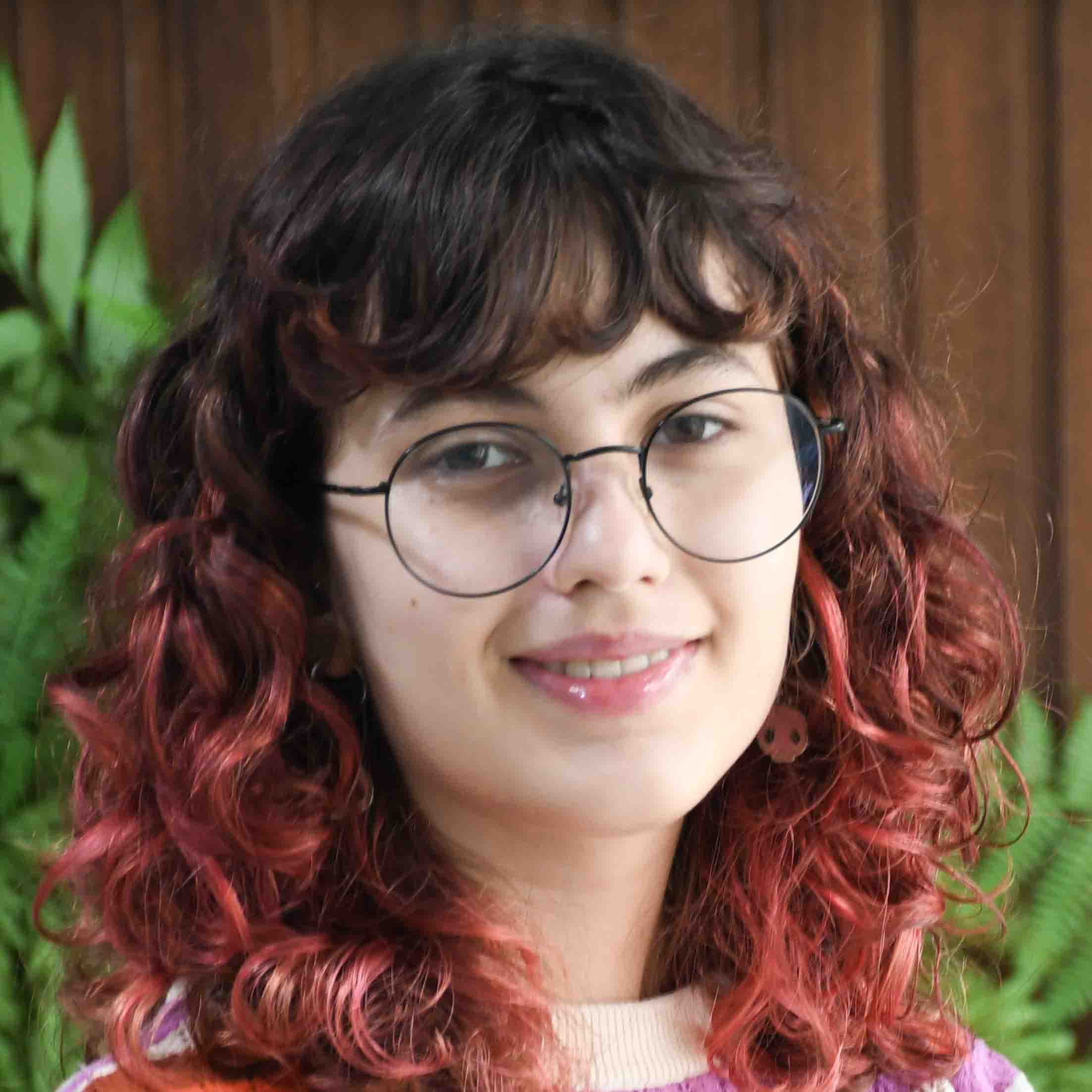 Camille, aluna do curso de ciência da computação, trajando um suéter lilás com detalhes claros. Ela está em um ambiente externo com fundo composto por madeira e plantas verdes, sugerindo um espaço natural ou decorativo. A iluminação natural realça os cabelos ondulados com tons avermelhados, criando um visual vibrante e descontraído.