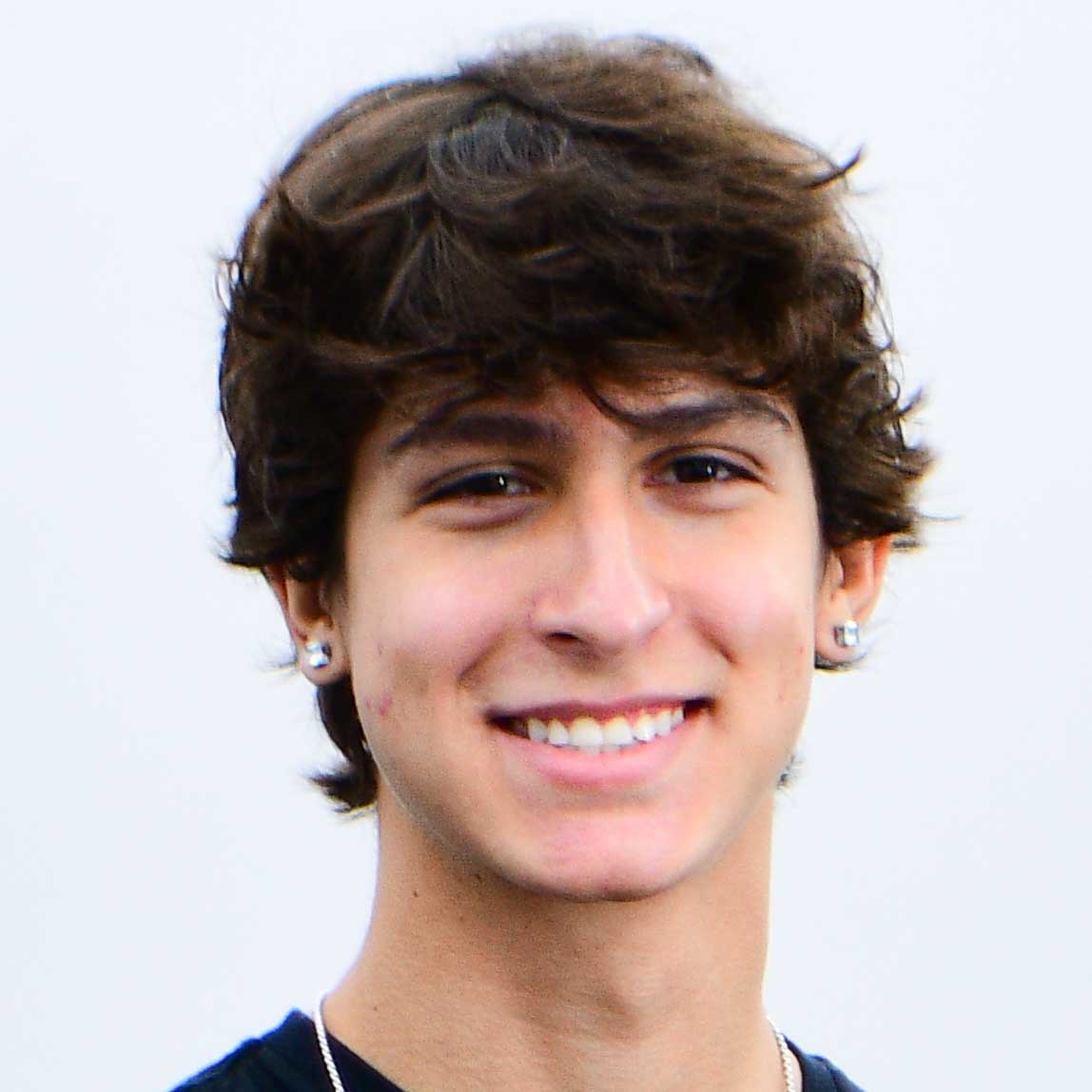 Filippo, aluno do curso de ciência da computação, trajando uma camiseta preta e um colar prateado. Ele está em um ambiente externo com fundo branco liso, sugerindo um dia claro ou um espaço neutro ao ar livre. A iluminação natural destaca os detalhes do cabelo castanho e o contraste entre a roupa escura e o fundo claro, criando uma composição simples e equilibrada.