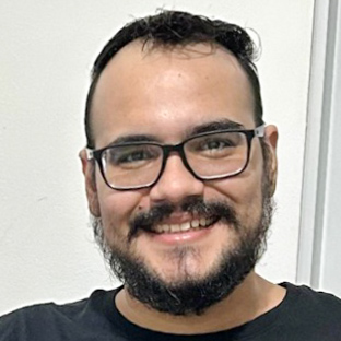 Irineu, egresso do curso de ciência da computação, trajando uma camiseta preta. Ele está em um ambiente interno com fundo branco e detalhes discretos de uma parede lisa, sugerindo um espaço formal ou acadêmico. A iluminação uniforme ressalta a simplicidade da composição, destacando o contexto profissional e acadêmico da imagem.