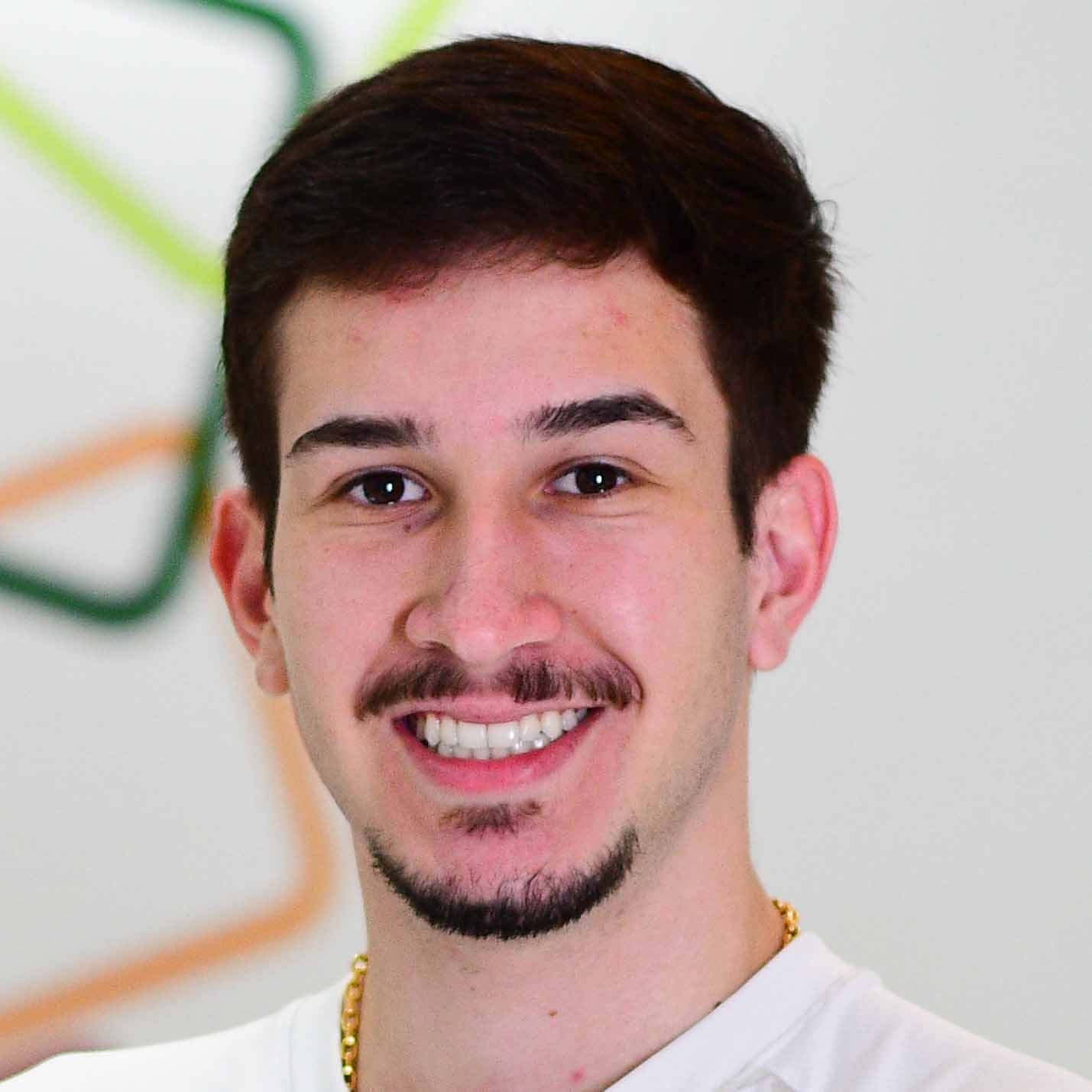 Pedro, aluno do curso de ciência da computação, trajando uma camiseta branca. Ele está em um ambiente interno com fundo branco decorado por linhas curvas coloridas em tons de verde e laranja, remetendo a um espaço institucional ou tecnológico. A iluminação clara valoriza o contraste entre a vestimenta e os elementos do fundo, reforçando o caráter moderno e acadêmico da imagem.