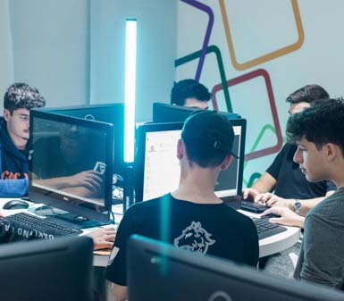 Grupo de estudantes de ciência da computação sentados ao redor de uma mesa redonda, cada um usando um computador de tela grande. Um tubo de luz vertical azul ilumina o centro da mesa, criando um efeito moderno. No fundo, a parede é decorada com linhas geométricas coloridas, transmitindo um ambiente inovador e tecnológico. Os alunos usam roupas escuras e estão concentrados nas telas, em clima de trabalho em equipe.