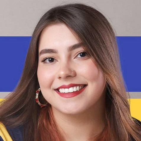 Gabriele, egressa de Ciências Biológicas - Licenciatura, posando em um ambiente com um fundo que remete à bandeira do Brasil. Ela tem cabelos castanhos claros e usa brincos grandes, com um sorriso largo.