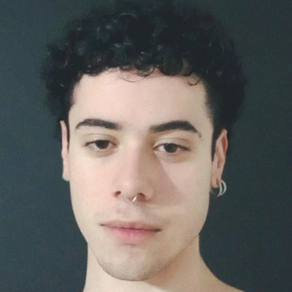 Nicholas, aluno de Ciências Biológicas - Licenciatura, posando em um ambiente interno com um fundo escuro. Ele tem cabelos curtos e cacheados, com piercings no nariz e na orelha, e uma expressão neutra.