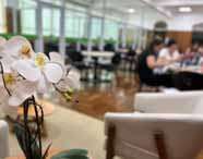 Ambiente interno aconchegante na Unoeste, com destaque para orquídeas brancas em primeiro plano. Ao fundo, mesas, cadeiras e grupos de estudantes reunidos, interagindo e estudando em um espaço confortável e iluminado, típico de áreas comuns universitárias.