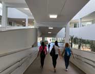 Três estudantes do curso de Enfermagem caminham por um corredor interno moderno e iluminado do campus da Unoeste em Jaú. Elas usam mochilas coloridas e roupas casuais, enquanto ao fundo há paredes brancas, janelas amplas, plantas decorativas e luz natural entrando pelas portas de vidro.