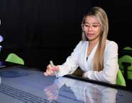 Aluna do curso de Enfermagem da Unoeste Jaú interage com mesa digital usando caneta touch, em sala escura com cadeiras verdes e equipamentos de realidade virtual ao fundo. Ela veste jaleco branco e óculos, concentrada na tela iluminada à sua frente.