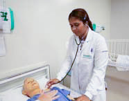 Aluna de Enfermagem em jaleco branco auscultando o tórax de um manequim de paciente com estetoscópio em laboratório, observada por outro aluno.