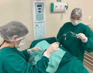 Duas alunas de Enfermagem da Unoeste Jaú, com avental verde, touca e luvas, realizam atividade prática em laboratório. Uma manipula seringa conectada a equipo, enquanto a outra auxilia próximo ao paciente coberto por campo cirúrgico. Ao fundo, painel de controle hospitalar