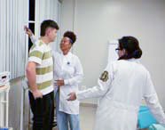 Três alunas de Enfermagem, uma delas medindo a altura de um jovem, enquanto as outras observam, em um ambiente de consultório ou laboratório.