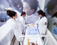 Três alunas de Enfermagem em jalecos brancos, incluindo uma professora, realizando uma aula prática em laboratório, examinando um manequim de bebê em um berço.