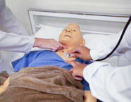 Duas alunas ou profissionais de saúde em jalecos brancos auscultando e palpando o tórax de um manequim de paciente idoso deitado em uma cama de hospital.