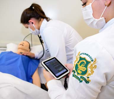 Duas estudantes do curso de Enfermagem da Unoeste Jaú realizam simulação prática em laboratório. Uma delas ausculta o manequim deitado na maca, coberto por avental azul, enquanto a outra registra informações em um tablet. Ambas usam jaleco branco com brasão do curso e máscara facial, demonstrando ambiente moderno de ensino.