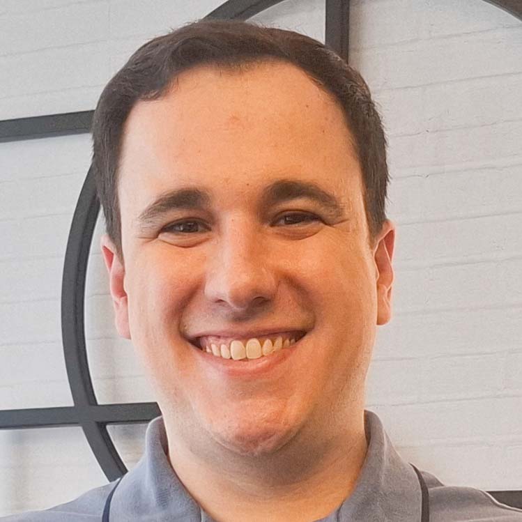 Vinicius, egresso do curso de análise e desenvolvimento de sistemas, trajando uma camisa cinza. Ele está em um ambiente interno com fundo composto por uma parede branca com detalhes geométricos pretos, sugerindo um espaço contemporâneo ou corporativo. A iluminação clara destaca a neutralidade da composição, transmitindo um ar profissional e elegante.