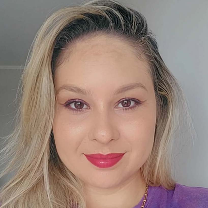 Amanda, aluna do curso de psicologia, trajando uma camiseta roxa. Ela está em um ambiente interno com fundo claro e neutro, sugerindo um espaço residencial ou acadêmico. A iluminação suave destaca os detalhes do cabelo loiro e a tonalidade da roupa, criando um visual harmônico e acolhedor.