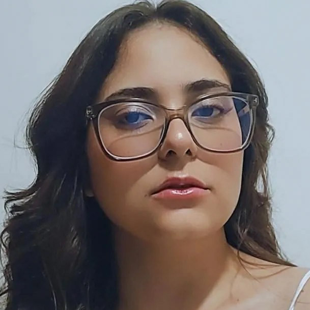 Isabele, aluna do curso de pedagogia, trajando uma blusa branca de alças finas. Ela está em um ambiente interno com fundo claro e neutro, sugerindo um espaço residencial ou acadêmico. A iluminação suave destaca os detalhes do cabelo castanho escuro e os óculos de armação grossa, criando um visual harmônico e acolhedor.