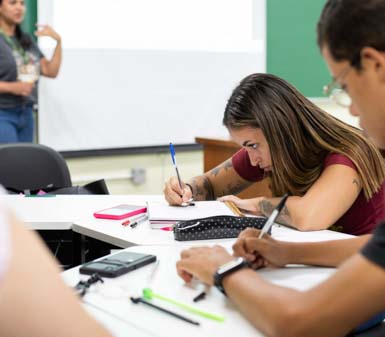 Alunos da Faculdade de História em Presidente Prudente concentrados em anotações durante aula, com professor ao fundo, representando o perfil profissional do curso focado em pesquisa e análise.