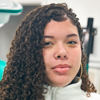 Imagem de Alicia, aluna do curso de enfermagem no campus Guarujá, vestindo um jaleco branco. Ela está posicionada em um ambiente acadêmico que aparenta ser uma sala de aula ou laboratório, com equipamentos médicos ao fundo, incluindo uma luminária odontológica. A iluminação da cena é clara, destacando o ambiente profissional e organizado.