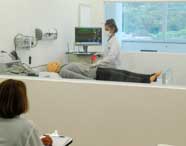 Laboratório de simulação do curso de Enfermagem da Unoeste Guarujá. Estudante de jaleco realiza procedimento em manequim deitado em maca, com monitor exibindo sinais vitais ao fundo. Equipamentos médicos conectados à parede e janela ampla com vista externa completam o ambiente moderno.