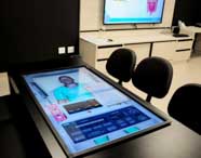 Laboratório de realidade virtual do curso de Enfermagem da Unoeste Guarujá, com mesa digital interativa exibindo imagem de paciente virtual e dados clínicos. Cadeiras pretas ao redor e tela grande na parede reforçam o uso de tecnologia avançada para ensino prático e simulações clínicas.