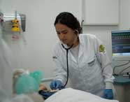 Aluna de Enfermagem em jaleco branco e luvas azuis auscultando o tórax de um manequim de paciente com um estetoscópio, em um laboratório de simulação, com monitor médico ao fundo.