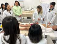 Grupo de estudantes do curso de Enfermagem da Unoeste Guarujá, reunidos ao redor de paciente simulado em maca. Alunos e professores usam jalecos brancos, alguns seguram papéis e outros manipulam equipamentos médicos, promovendo aprendizado prático em ambiente laboratorial