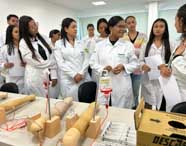 Turma do curso de Enfermagem da Unoeste Guarujá em laboratório, todos de jaleco branco. Alunos se reúnem em torno de mesa com simuladores de braços para punção venosa, bolsas de soro e materiais médicos, praticando técnicas essenciais para a formação profissional