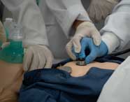 Close-up das mãos de alunos de Enfermagem em luvas, realizando ausculta e manuseando equipamentos em um manequim de paciente em atividade prática.