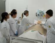 Alunos de Enfermagem em jalecos brancos praticando cuidados em um manequim de paciente em cama de hospital, com um aluno auscultando e outros observando, em ambiente de simulação.