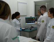 Alunos de Enfermagem em jalecos brancos observando e interagindo com um monitor que exibe sinais vitais e gráficos, em um laboratório de simulação hospitalar.