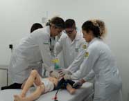 Quatro alunos de Enfermagem em jalecos brancos realizando um exercício prático de ventilação ou reanimação em um manequim de paciente, com um deles utilizando uma máscara.