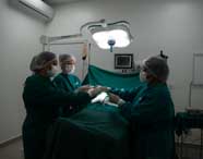 Três alunas de Enfermagem ou Medicina em vestimentas cirúrgicas (scrubs verdes, toucas e máscaras) realizando simulação de procedimento cirúrgico sob iluminação focada.