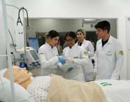Grupo de alunos de Enfermagem em jalecos brancos ao redor de um manequim de paciente em uma cama de hospital, praticando procedimentos como administração de soro.