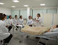 Professora de Enfermagem em jaleco branco instruindo um grupo de alunos sentados em um laboratório de simulação hospitalar, com um manequim de paciente na cama.