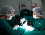 Grupo de alunos de Enfermagem em scrubs e equipamentos de proteção, realizando uma simulação de procedimento cirúrgico, com foco na área operatória.