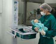Aluna de Enfermagem ou profissional de saúde em scrubs e touca, segurando um manequim de bebê recém-nascido ao lado de uma incubadora em um laboratório de simulação.