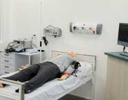 Leito hospitalar equipado para simulação realista no curso de Enfermagem da Unoeste Guarujá. Manequim adulto deitado, monitor multiparamétrico ligado, aparelhos médicos na parede e mesa lateral com desfibrilador e outros equipamentos, criando ambiente fiel ao atendimento hospitalar.