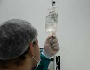 Close-up de um aluno de Enfermagem ou profissional de saúde em scrubs, touca e máscara, segurando um saco de soro intravenoso, ajustando o fluxo ou preparando a administração.
